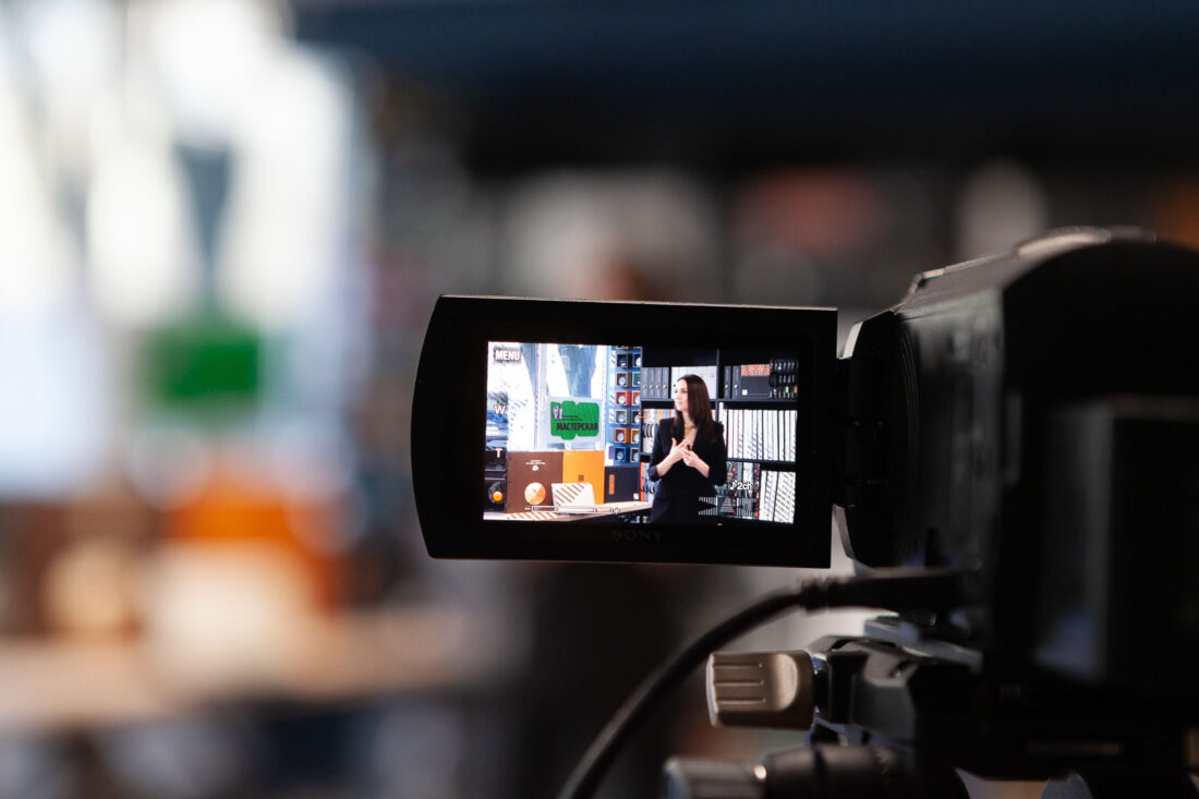 Съёмка лекции о дизайне интерьера: кадр через экран видеокамеры, на котором видно выступление спикера на фоне стеллажей с образцами материалов в шоуруме.