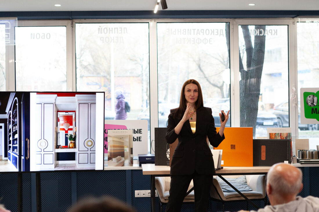 Лекция о современном интерьере в дизайн‑центре Ампир Декор на Автозаводской: спикер в тёмном костюме выступает перед аудиторией на фоне большого экрана с фотографиями реализованного проекта и стеллажа с образцами отделочных материалов.