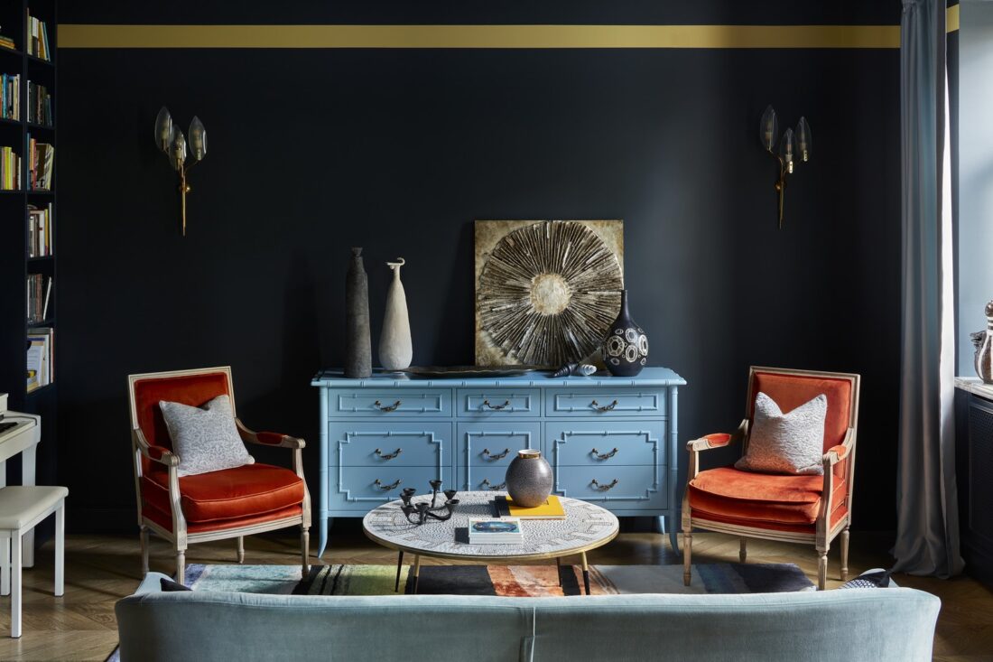 Гостиная с тёмно‑синими стенами и золотой полосой под потолком, винтажным голубым комодом, двумя оранжевыми креслами, круглым журнальным столиком и декоративными вазами и панно в эклектичном интерьере.