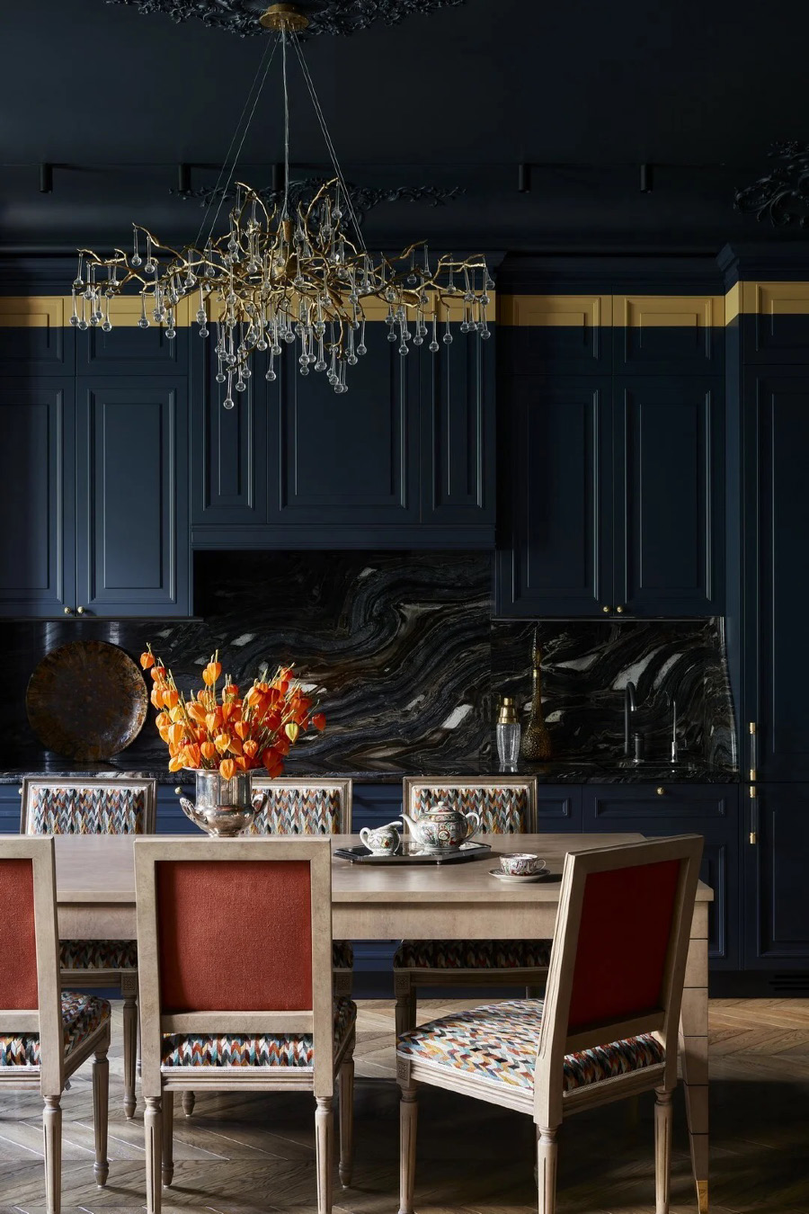 Столовая‑кухня с тёмно‑синим классическим гарнитуром и мраморным фартуком, деревянным обеденным столом и стульями с оранжевыми спинками, крупной люстрой с подвесками и букетом оранжевых цветов в центре.