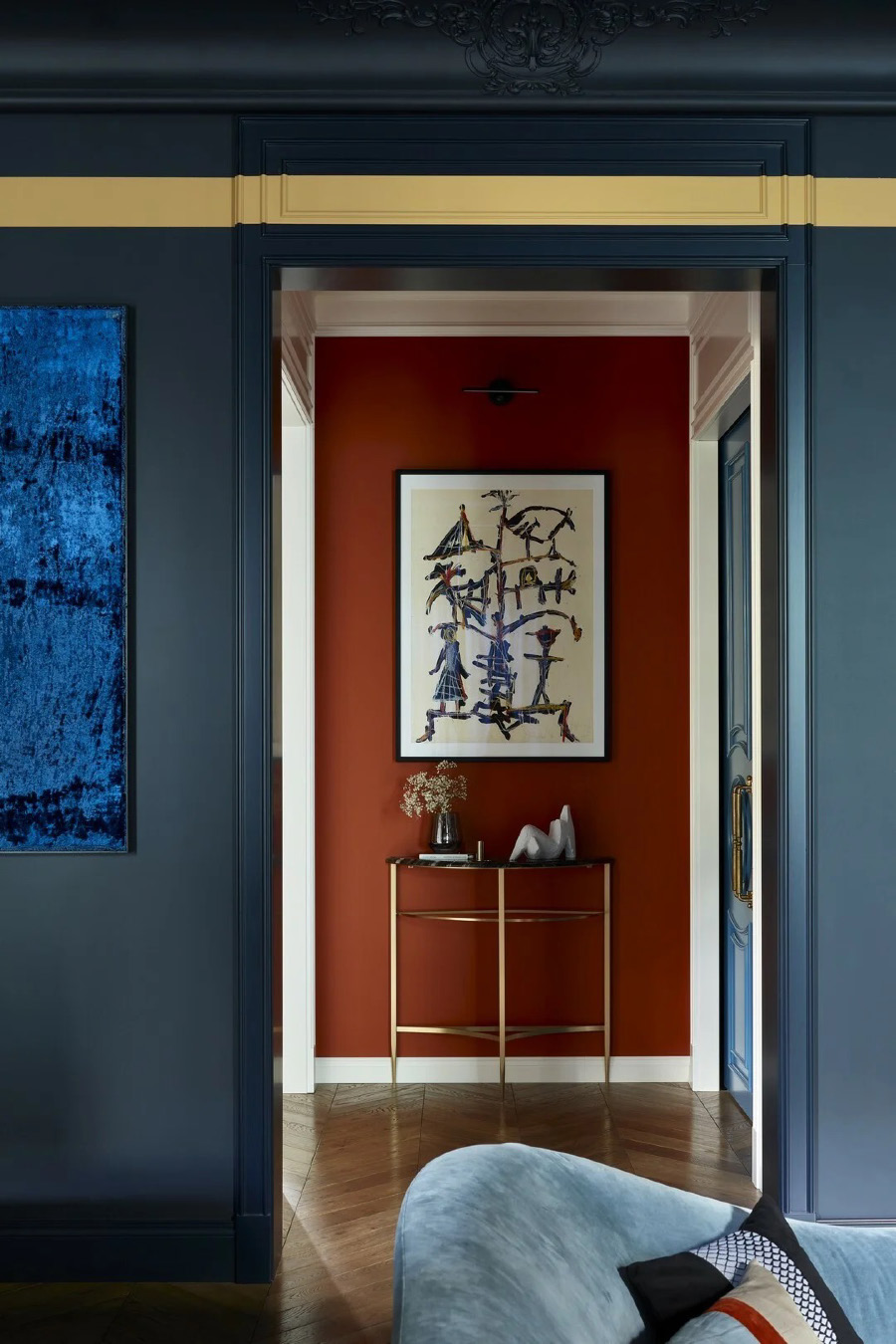 Вид из гостиной в коридор с терракотовой стеной, золотой консолью и графичной картиной, обрамлённый тёмно‑синими панелями с золотой полосой и частью голубого дивана на переднем плане.