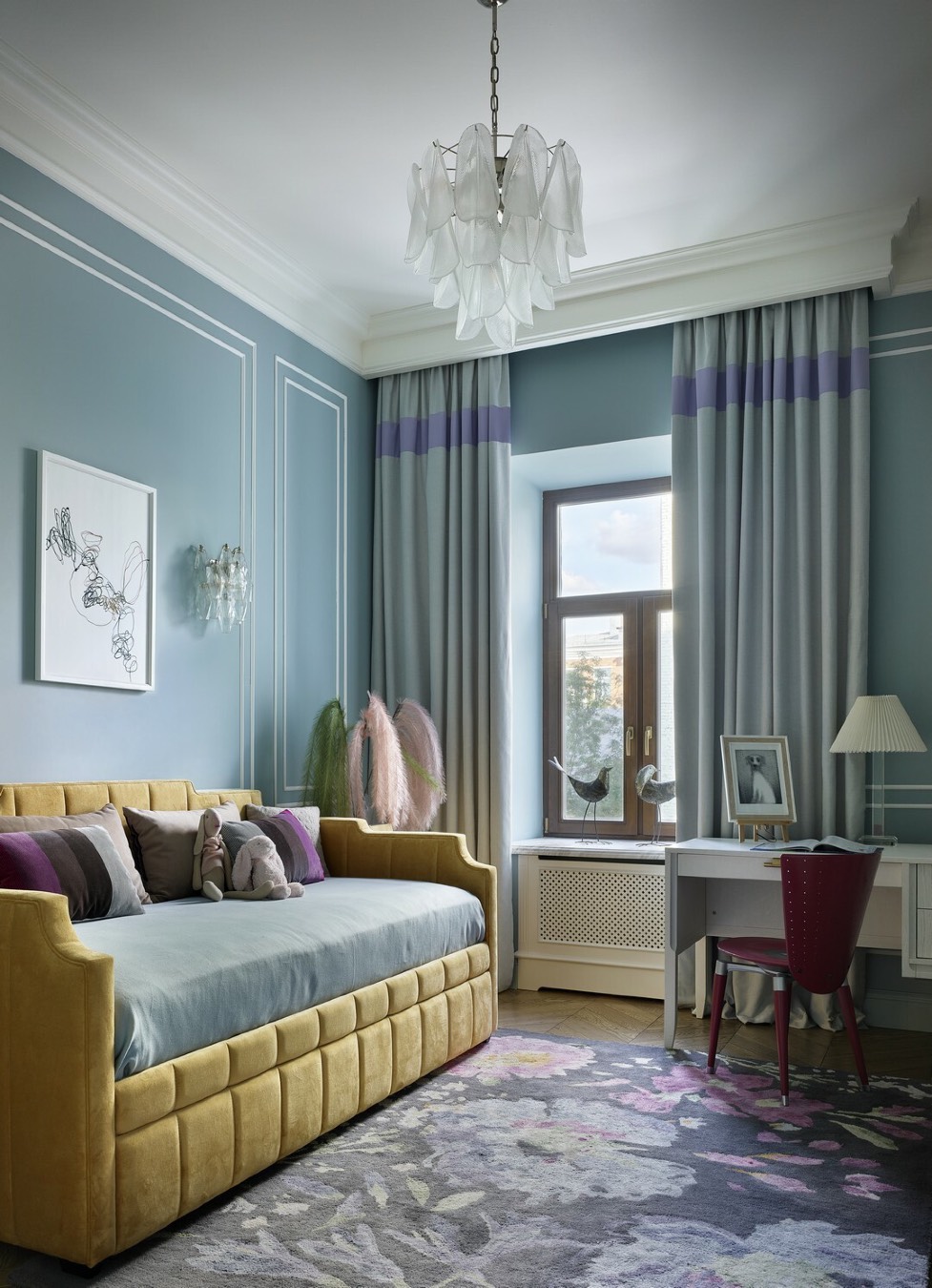 Детская комната в голубых тонах с жёлтым мягким диваном‑кроватью, серо‑лиловыми шторами, письменным столом у окна и ковром с крупным цветочным рисунком.