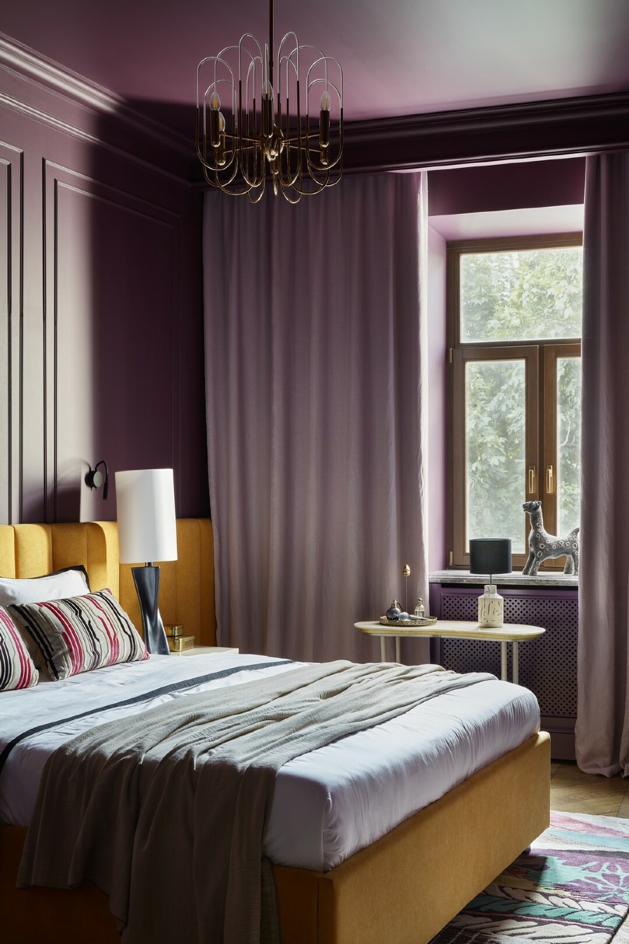 Спальня в сложных фиолетовых тонах с однотонными стенами и потолком, жёлтым мягким изголовьем кровати, светло‑серыми текстилем и шторами, круглым прикроватным столиком и окном с видом на зелень.