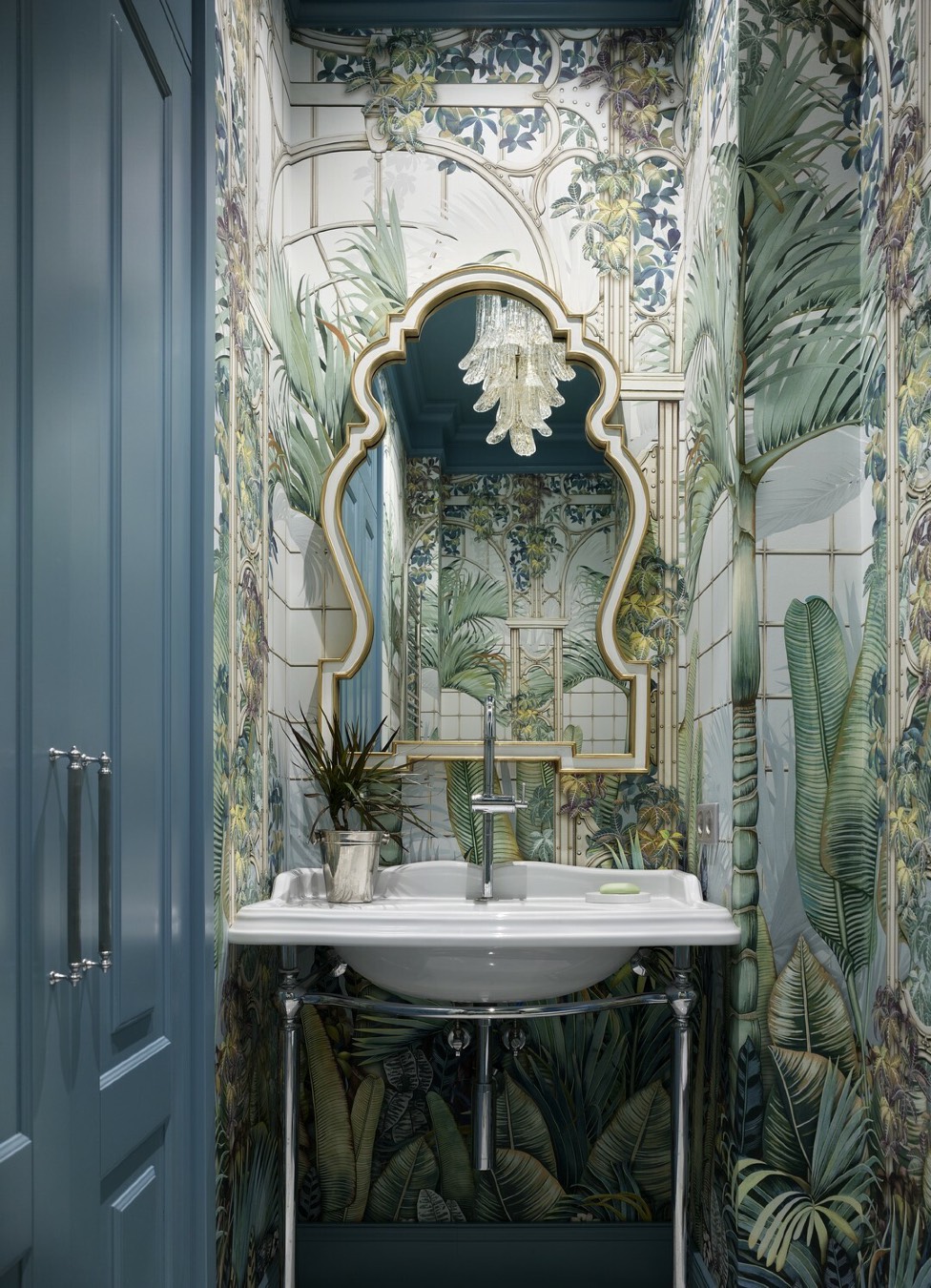 Гостевой санузел с раковиной на хромированных ножках, стенами в тропических обоях, фигурным зеркалом в золотой раме и хрустальной люстрой на фоне голубых панелей.