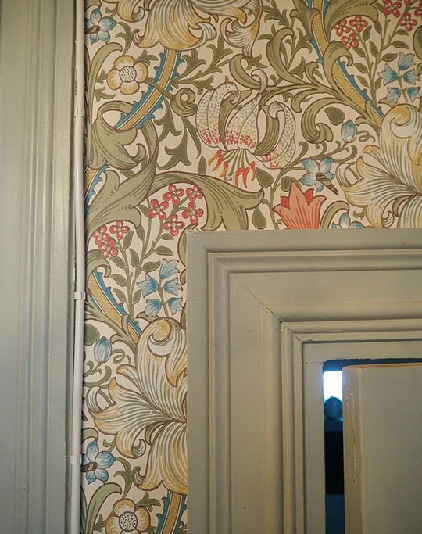 Фрагмент стены с обоями в стиле Уильяма Морриса: плотный растительный орнамент с крупными листьями и цветами в зелёных, кремовых, голубых и коралловых оттенках. Справа видно часть дверного проёма с массивным наличником, выкрашенным в светло‑серо‑зелёный цвет.