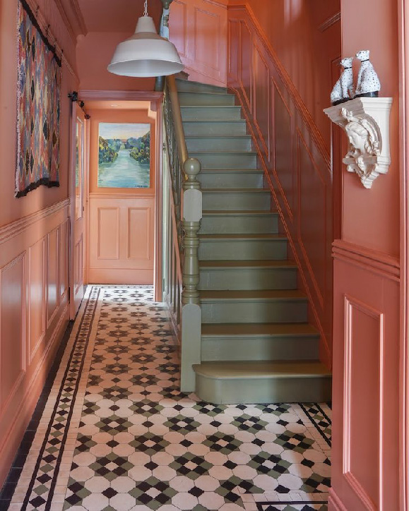 Яркий коридор с лестницей: стены и двери выкрашены в тёплый кораллово‑розовый цвет, пол выложен плиткой с чёрно‑белым геометрическим узором. В центре — лестница оливково‑зелёного цвета с перилами, наверху висит белый подвесной светильник. На стенах — картина с пейзажем в конце коридора и текстильная панель слева, справа на консоли стоят декоративные фигурки.