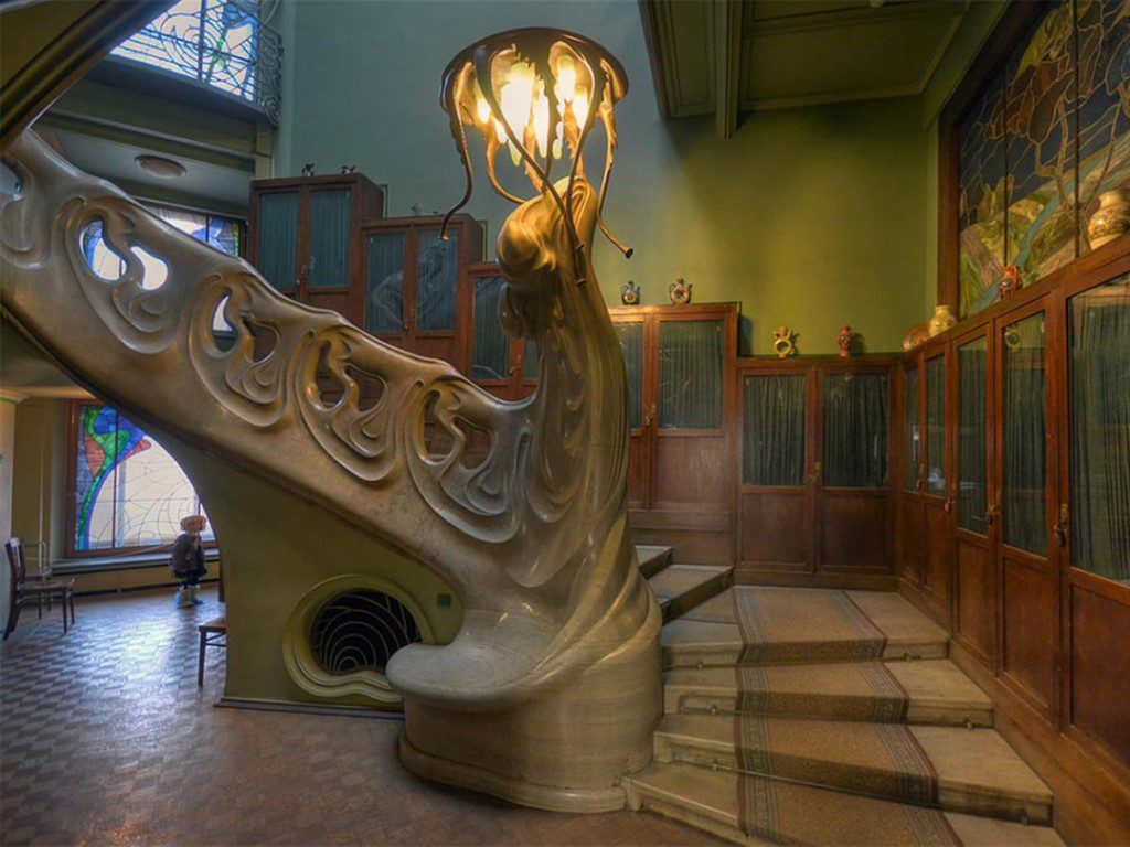 Интерьер в стиле модерн с монументальной волнообразной лестницей и скульптурным светильником, плавно перетекающим в перила, витражными окнами и деревянными шкафами с декоративными вставками.