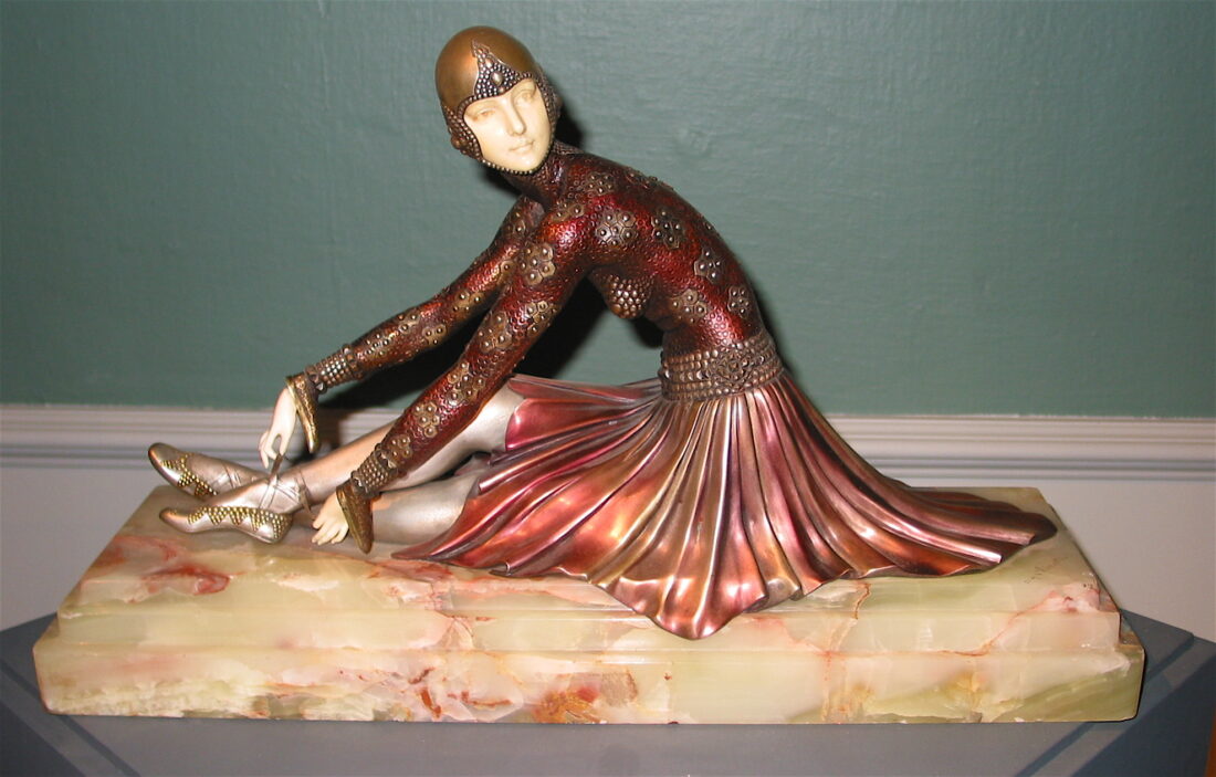 Скульптура в стиле ар-деко: сидящая танцовщица в шлеме и украшенном костюме с длинной металлически переливающейся юбкой, расположенная на светлом каменном постаменте.