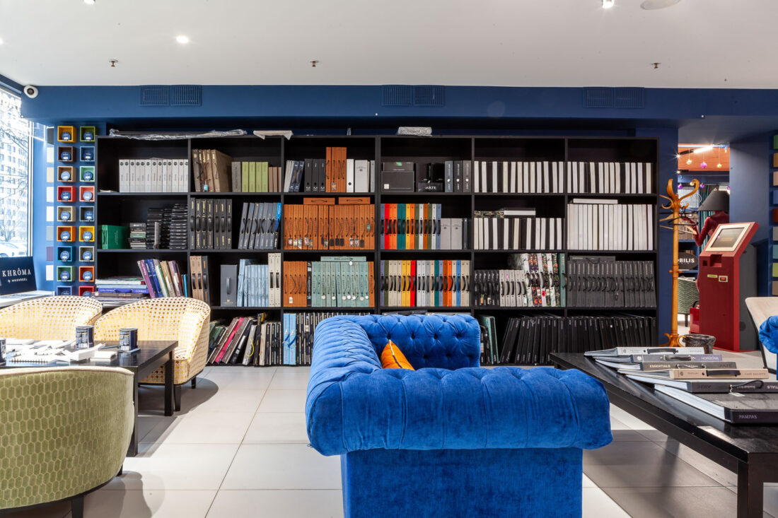 Зона для работы и встреч в дизайн‑центре Ампир Декор на Автозаводской с синим диваном и высоким стеллажом, заполненным каталогами обоев Arte, Omexco, Masureel