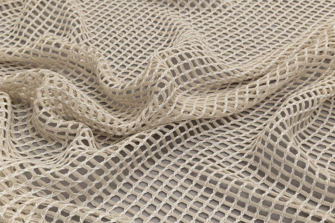 Крупный план бежевой тюль-сетки с крупным переплетением нитей, лежащей мягкими складками на светлом фоне.