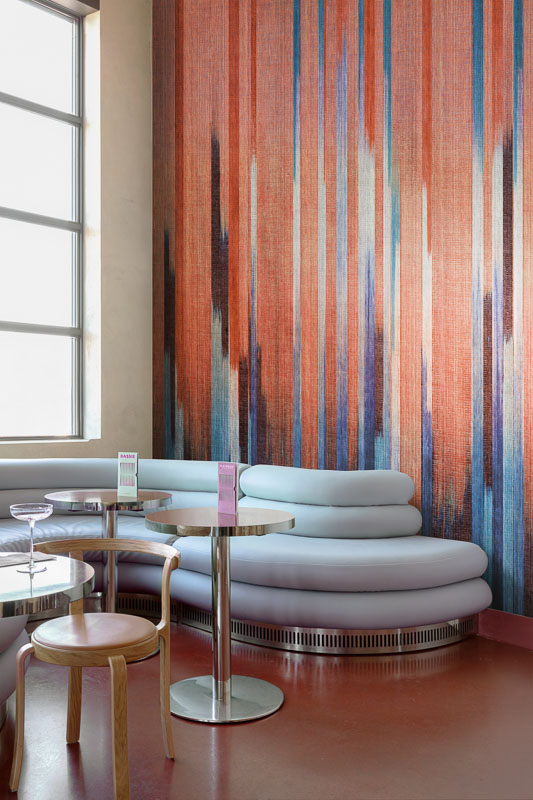 Интерьер зоны отдыха с мягким изогнутым диваном и круглыми столиками у стены, оформленной ярким декоративным настенным панно с вертикальным градиентным рисунком.