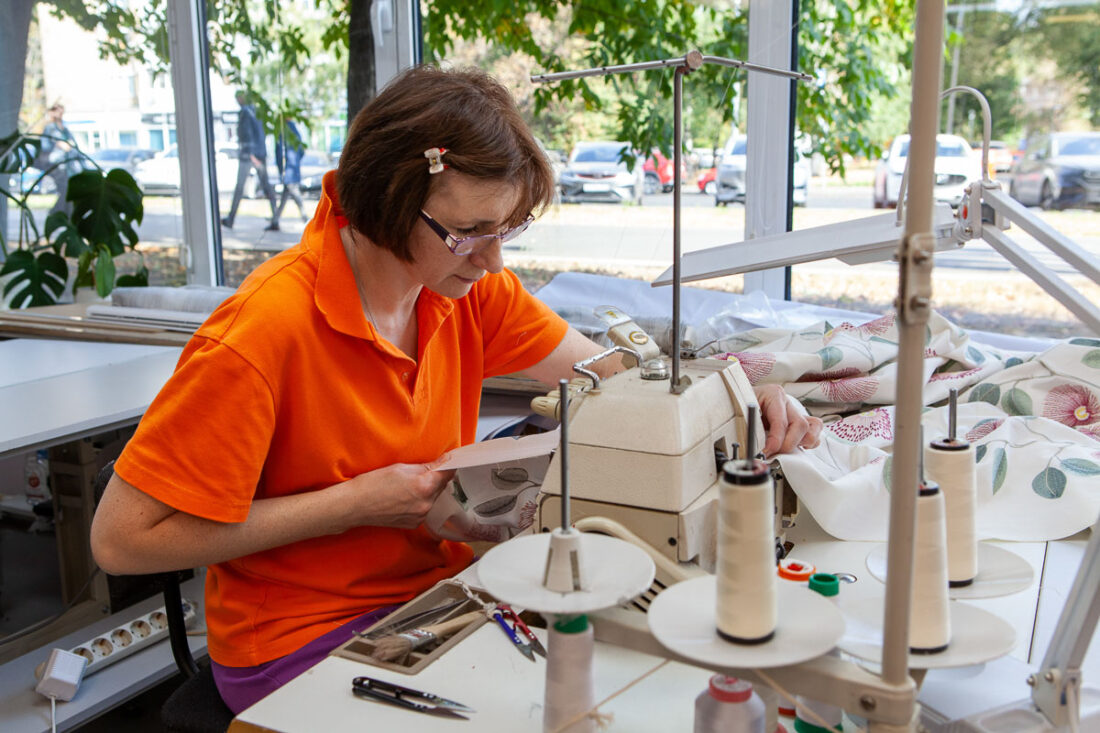 Швея за работой за промышленной швейной машиной, обрабатывает ткань с цветочным рисунком у панорамного окна — швейный цех Ампир Декор на Автозаводской.