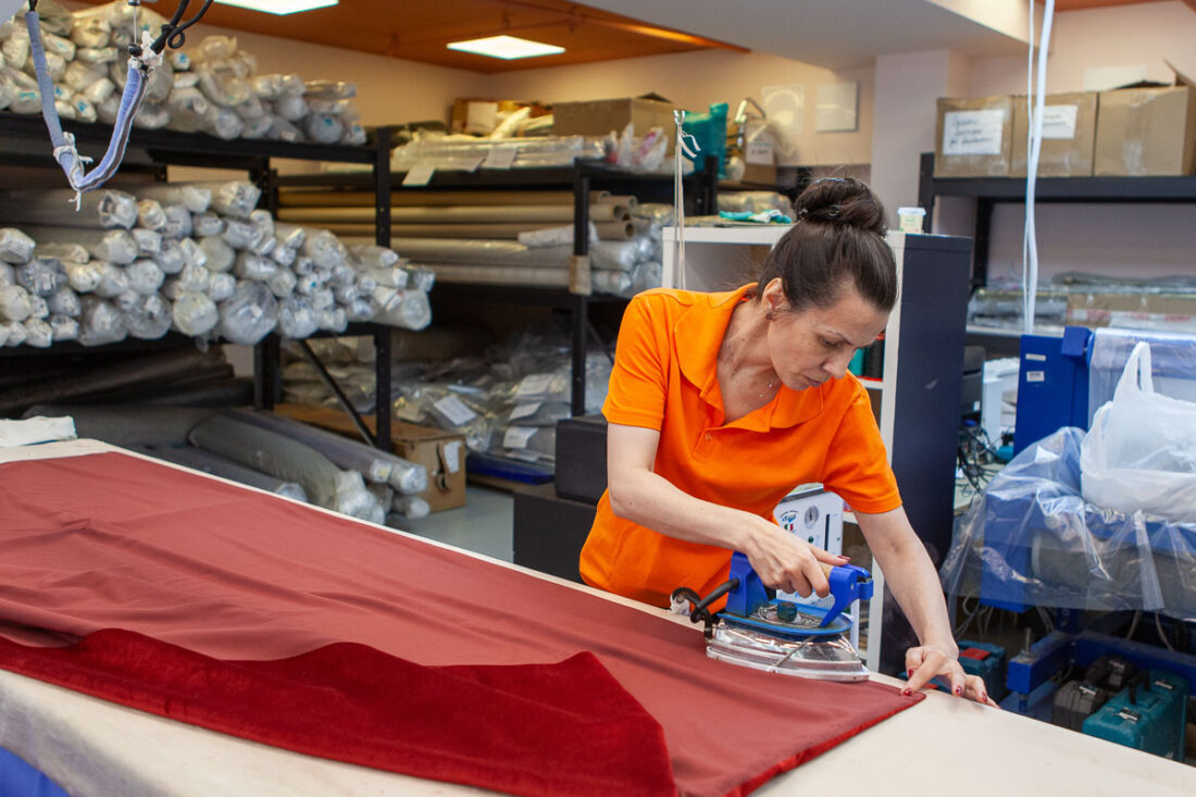 Швея в оранжевой форме отпаривает и утюжит бордовую портьерную ткань на рабочем столе, на фоне складских стеллажей — швейный цех Ампир Декор на Автозаводской.