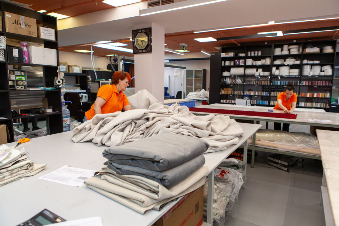 Две сотрудницы в оранжевой форме раскладывают и готовят к пошиву светлые портьеры и ткани на больших столах — швейный цех Ампир Декор на Автозаводской.