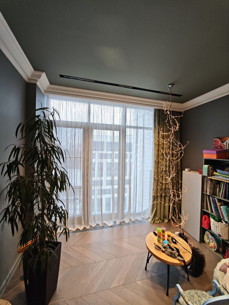 Детская комната с большой оконной стеной, оформленной тюлем-сеткой и зелёными портьерами с жаккардовым рисунком, деревянным полом, игровой зоной и стеллажом с книгами и игрушками.