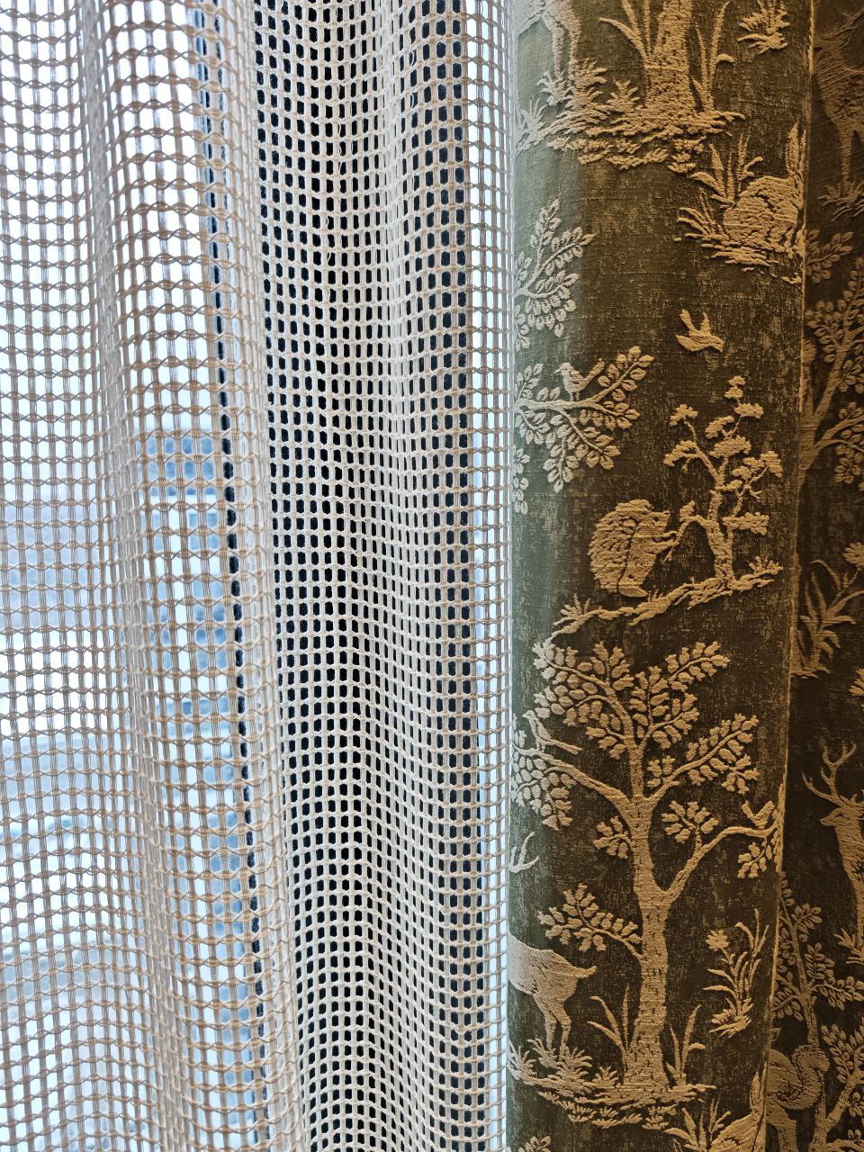 Крупный план светлой тюли-сетки на окне в сочетании с зелёной портьерой из бархата с жаккардовым анималистическим рисунком леса и животных.