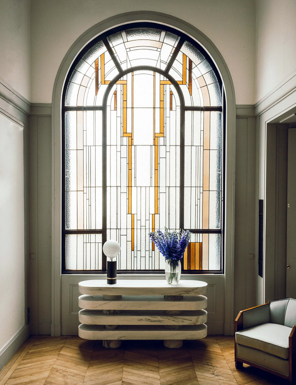 Холл в стиле ар-деко: большое арочное витражное окно с геометрическим узором, перед ним кремовая консоль с круглой лампой и вазой с синими цветами, справа светлое кресло.