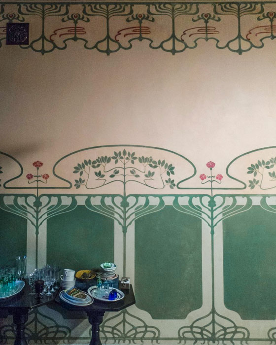 Стена в стиле модерн с зелёно-кремовым орнаментом: в нижней части крупные прямоугольные панели с плавными линиями, над ними декоративные ветви с листьями и розовыми цветами. Внизу слева стоят два тёмных столика, заставленные посудой и стеклянной посудой.