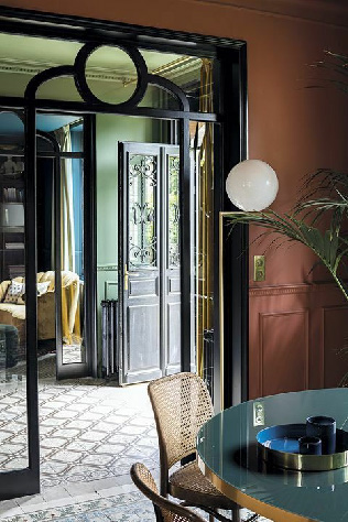 Современный интерьер с мотивами модерна: анфилада комнат, декоративные двери с изогнутыми линиями, чёрные порталы, зелёные и терракотовые стены, узорный пол. В кадре круглый стол, плетёный стул и мягкий рассеянный свет.