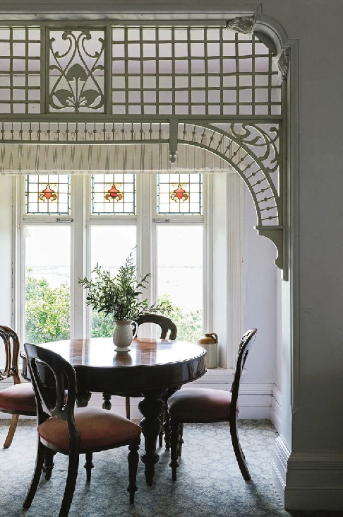 Светлая столовая в стиле модерн с резной декоративной перегородкой, витражными окнами, круглым деревянным столом и изящными стульями, оформленная в спокойной природной гамме.