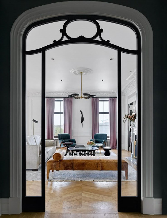 Элегантная современная гостиная в светлых тонах с высокими окнами, лиловыми шторами, изумрудными креслами и минималистичным декором, видимая через черную металло‑стеклянную арку.